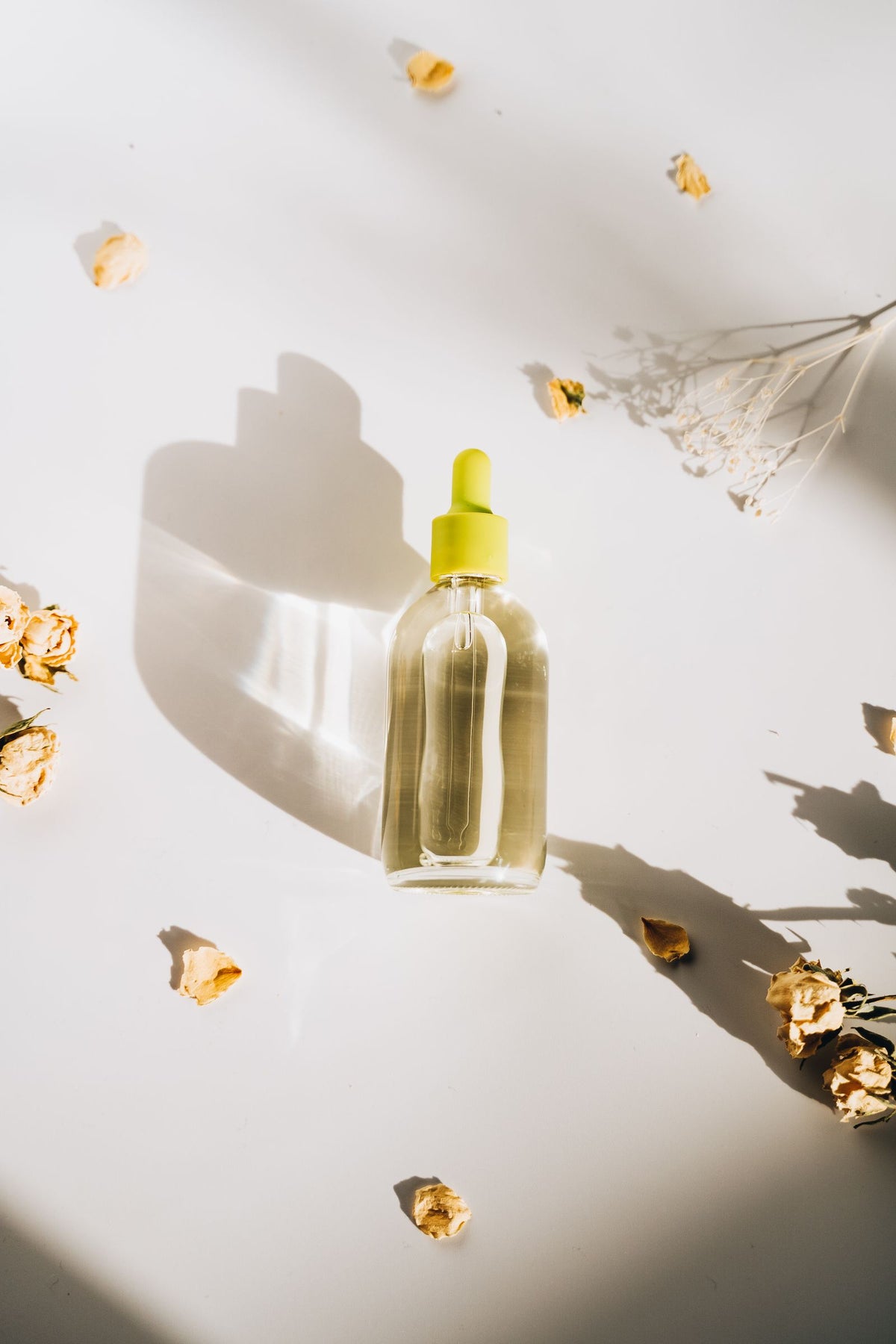 Frasco de sérum transparente sobre superfície branca, cercado por flores secas