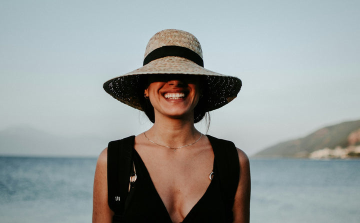 Mulher sorrindo com chapéu de abas largas à beira-mar